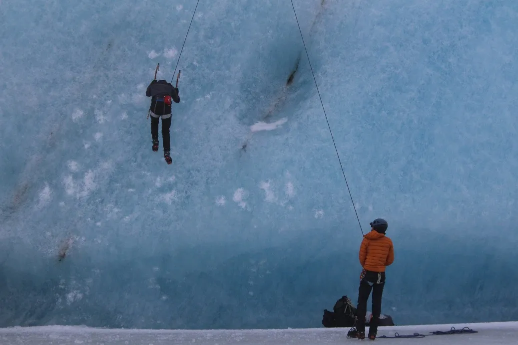 A man climbs a vertical ice wall while his friend belays him