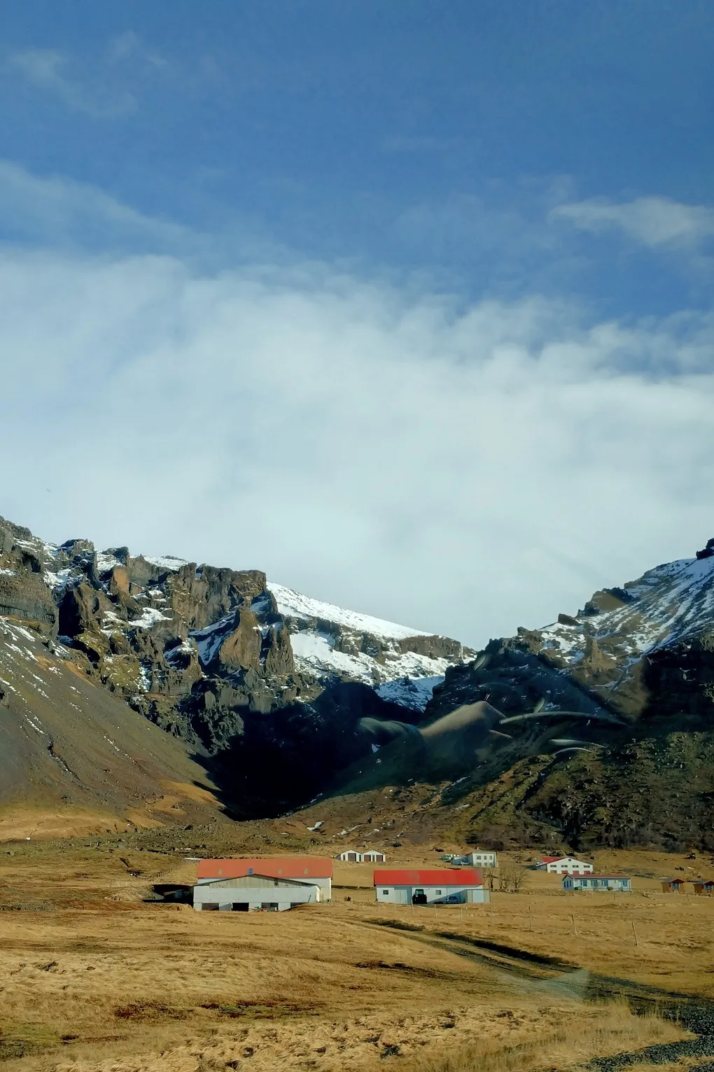 A cluster of houses and barns, all with grey walls and red roofs with the mountains looming behind